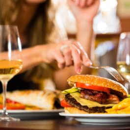 O femeie care se află la o masă și mănâncă dintr-un burger care stă alături de două pavare de vin alb, fiind produse care se află pe lista de alimente și băuturi pe care ar trebui să le eviți
