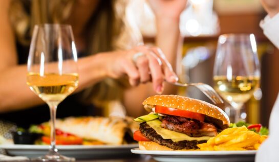 O femeie care se află la o masă și mănâncă dintr-un burger care stă alături de două pavare de vin alb, fiind produse care se află pe lista de alimente și băuturi pe care ar trebui să le eviți
