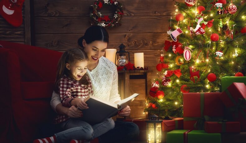 O mamă alături de fiica sa lângă un brad frumos împodobit în timp ce îi citește una din cele opt cărți pentru copii cu tematică de Crăciun