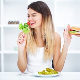 O femeie frumoasă blondă care stă în fața unei farfurii pe care se află un centimetru, în timp ce ține în mână de salată și împinge un burger