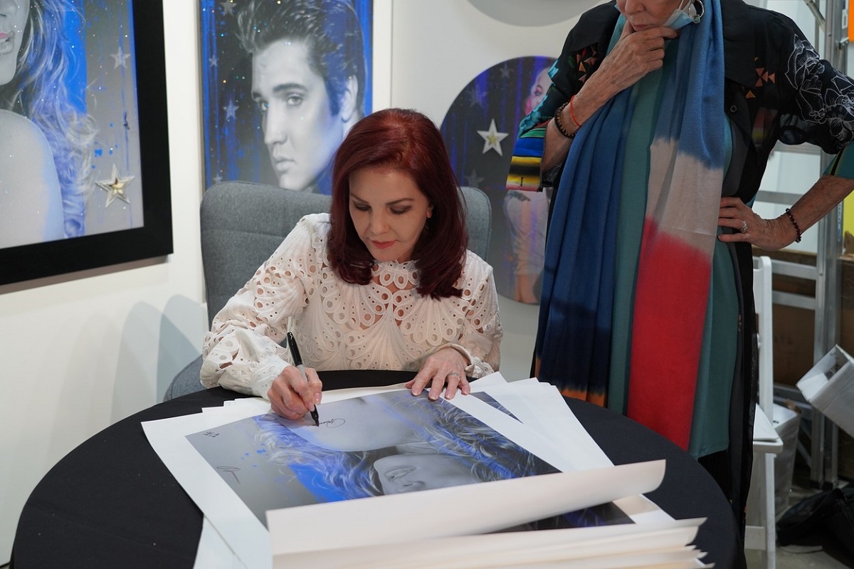 Priscilla Presley la o masă în timp ce semnează un autoportret realizat de Adam Rote