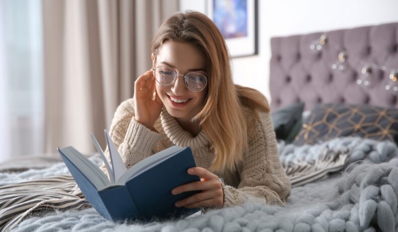 O femeie frumoasă, blondă, care stă în pat și poartă ochelari în timp ce citește una din cele cinci crăți de dezvoltare personală pentru femei