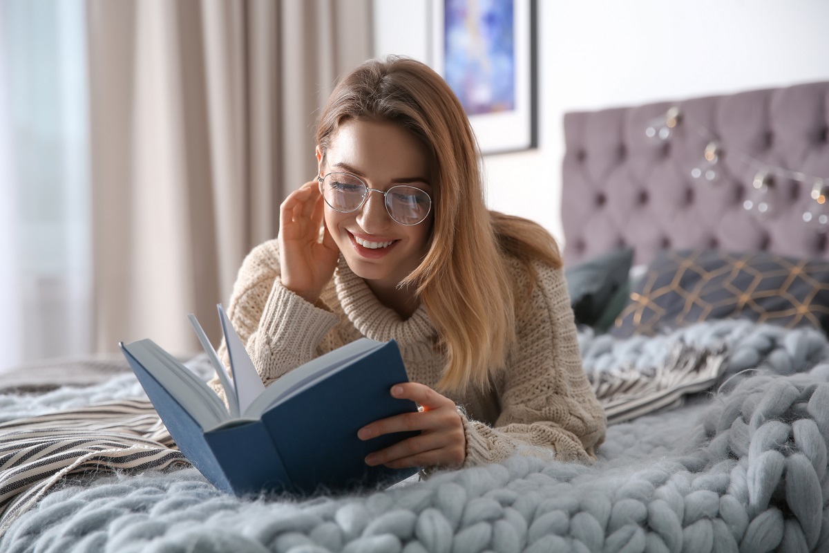 O femeie frumoasă, blondă, care stă în pat și poartă ochelari în timp ce citește una din cele cinci crăți de dezvoltare personală pentru femei
