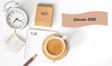 Un birou alb pe care se află o cana de cafea, un creion și un ces alături de o agendă și un calendar cu zile libere în anul 2022