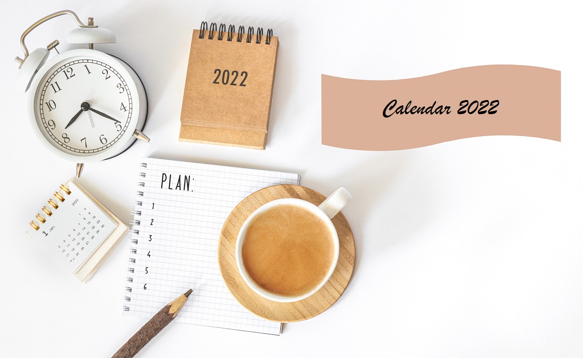 Un birou alb pe care se află o cana de cafea, un creion și un ces alături de o agendă și un calendar cu zile libere în anul 2022