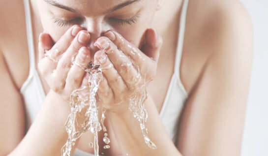 o femeie îmbrăcată într-u maiou alb, se spală corect pe față cu apă rece.
