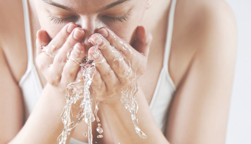 o femeie îmbrăcată într-u maiou alb, se spală corect pe față cu apă rece.