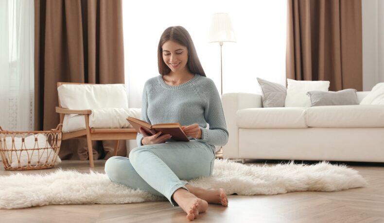 O femeie frumoasă care stă pe jos în fața unei canapele și citește dintr-o carte de educație financiară