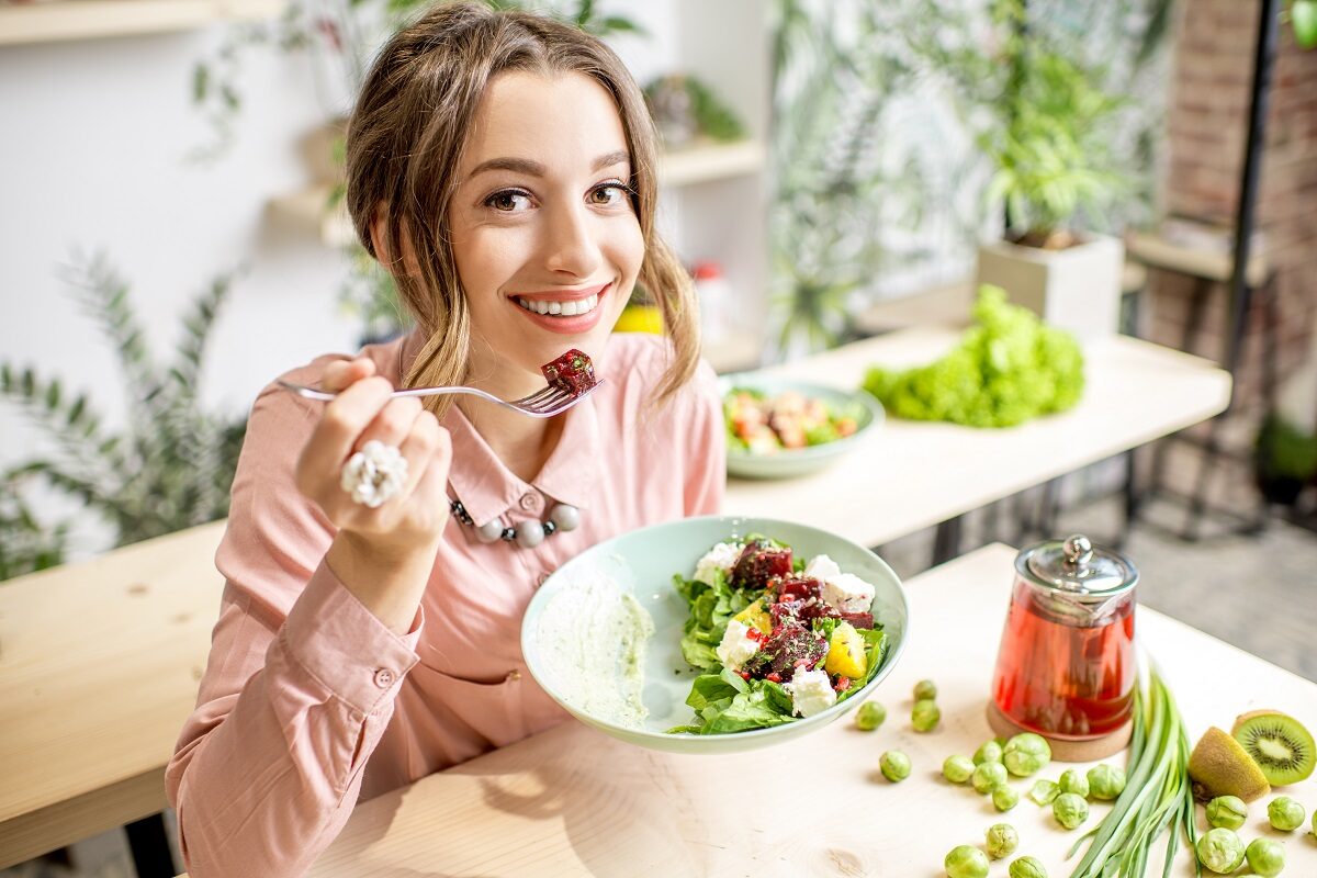 O femeie frumoasă care stă la masă și mănâncă dintr-o salată pentru a demonstra unul din cele cinci obiceiuri sănătoase care te ajută să pierzi în greutate