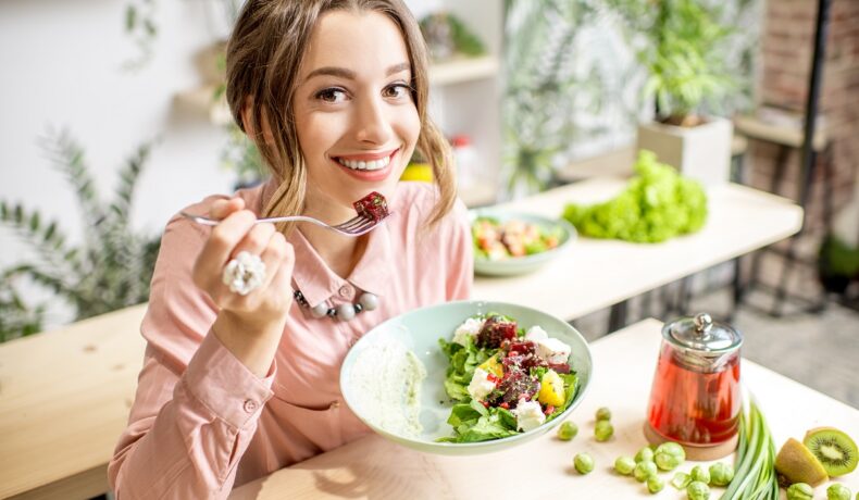 O femeie frumoasă care stă la masă și mănâncă dintr-o salată pentru a demonstra unul din cele cinci obiceiuri sănătoase care te ajută să pierzi în greutate