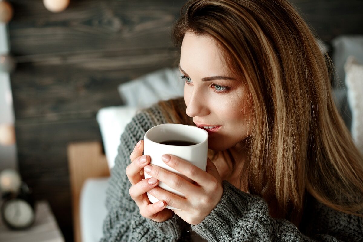 O femeie frumoasă care poartă un pulover gri și ține în mână o ceașcă de ceai negru, una din principalele băuturi care te ajută să adormi mai ușor