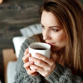 O femeie frumoasă care poartă un pulover gri și ține în mână o ceașcă de ceai negru, una din principalele băuturi care te ajută să adormi mai ușor