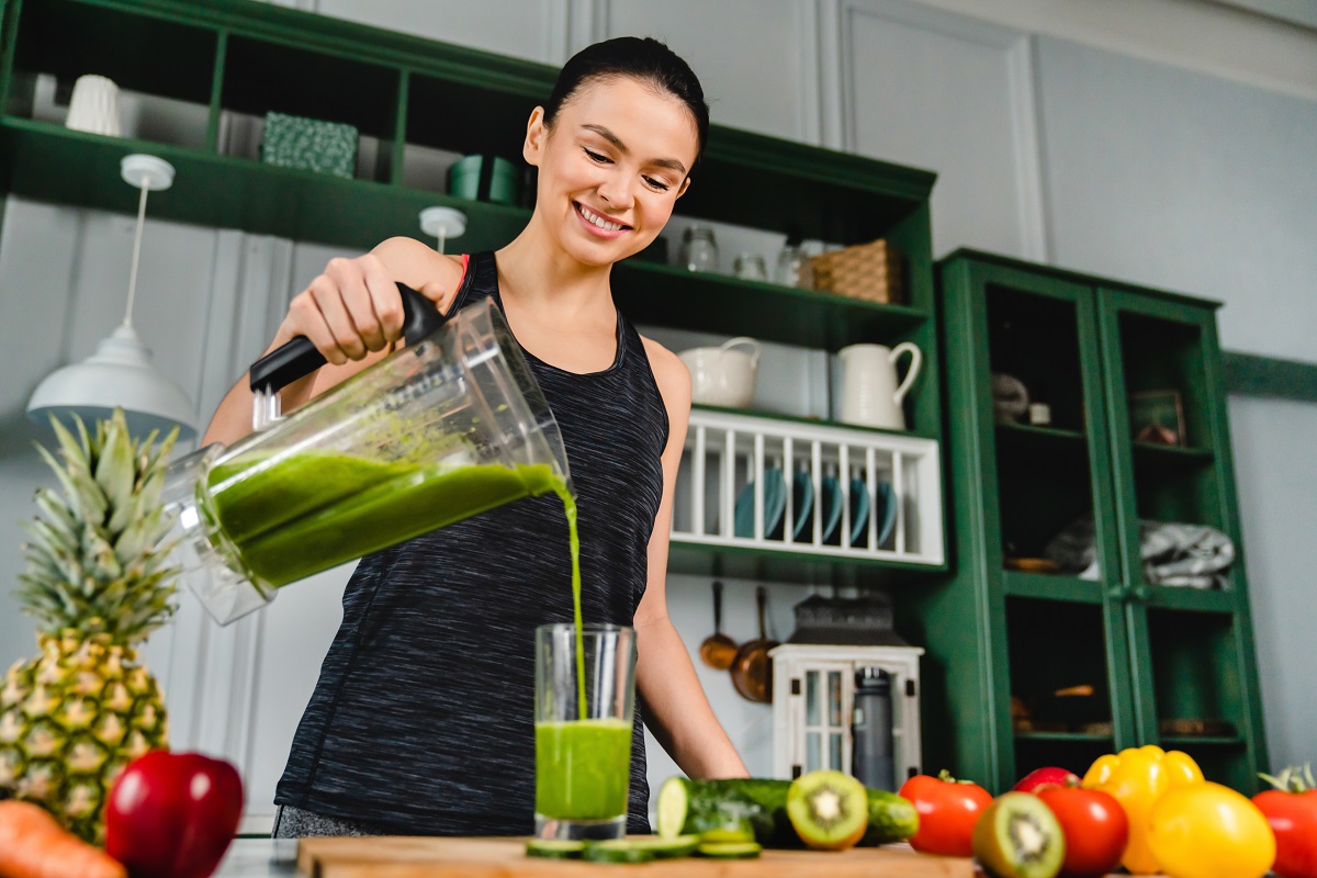 O femeie frumoasă într-o bucătărie în timp ce toarnă un suc verde din blender într-un pahar după ce a preparat unul din cele cinci tipuri de smoothie care previn îmbătrânirea
