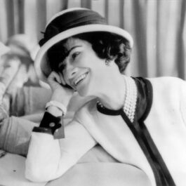 Coco Chanel într-un costum alb, cu pălărie, în timp ce zâmbește