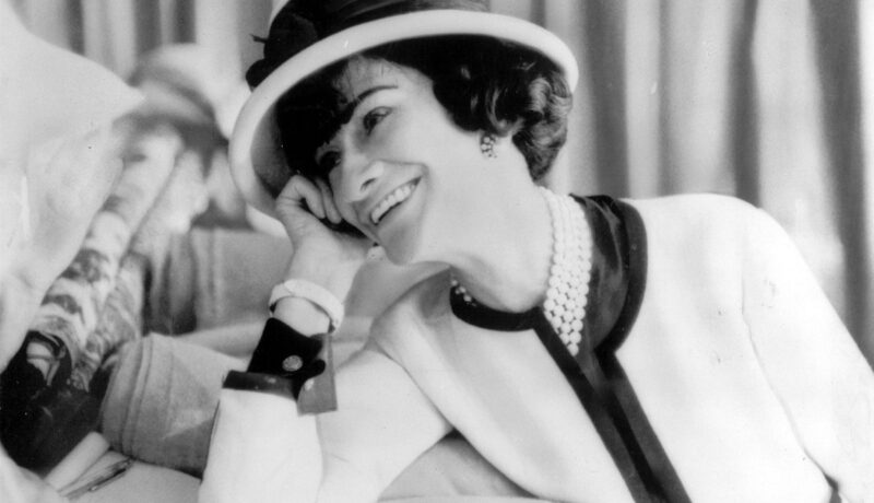 Coco Chanel într-un costum alb, cu pălărie, în timp ce zâmbește