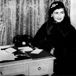 Coco Chanel într-un costum negru în timp ce stă în fața unui birou