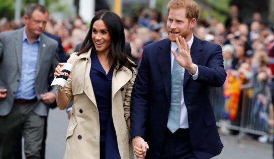 Meghan Markle și Prințul Harry în timp ce se țin de mână și salută publicul înc adrul unei întâlniri oficiale din Regatul Unit