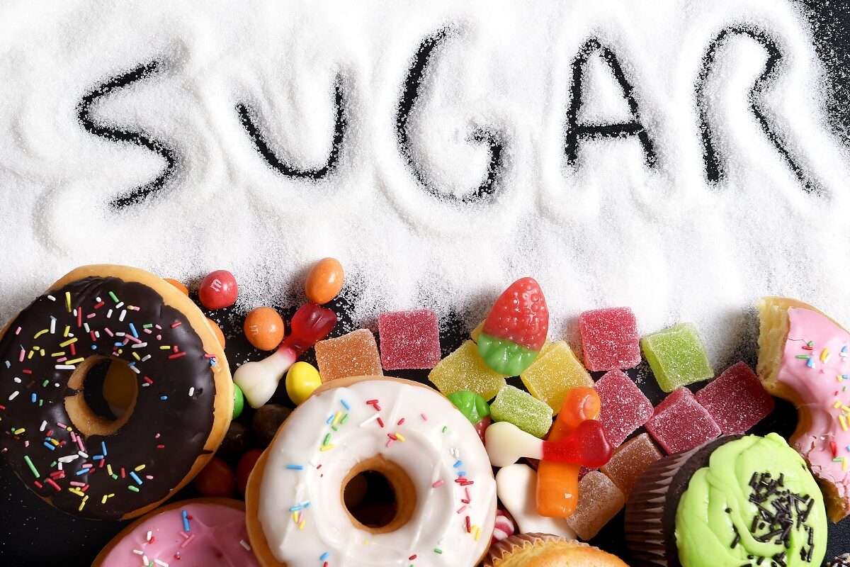 O masă pe care se află mai multe gogoși glazurate și zahăr în care este scris cuvântul sugar pentru a arăta care este ingredientul alimentar care îți poate afecta sănătatea
