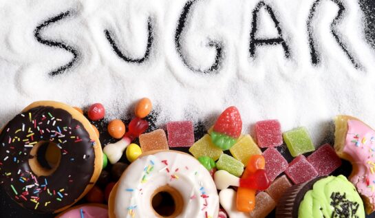 O masă pe care se află mai multe gogoși glazurate și zahăr în care este scris cuvântul sugar pentru a arăta care este ingredientul alimentar care îți poate afecta sănătatea