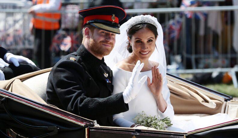 Prințul Harry alături de Meghan Markle îmbrăcați pentru ceremonia de nuntă din anul 2018 în timp ce fac cu mâna publicului dintr-o trăsură