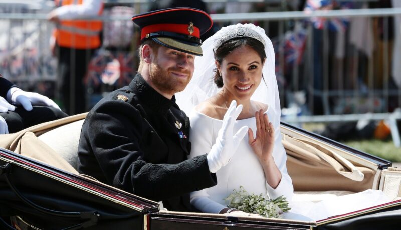 Prințul Harry alături de Meghan Markle îmbrăcați pentru ceremonia de nuntă din anul 2018 în timp ce fac cu mâna publicului dintr-o trăsură