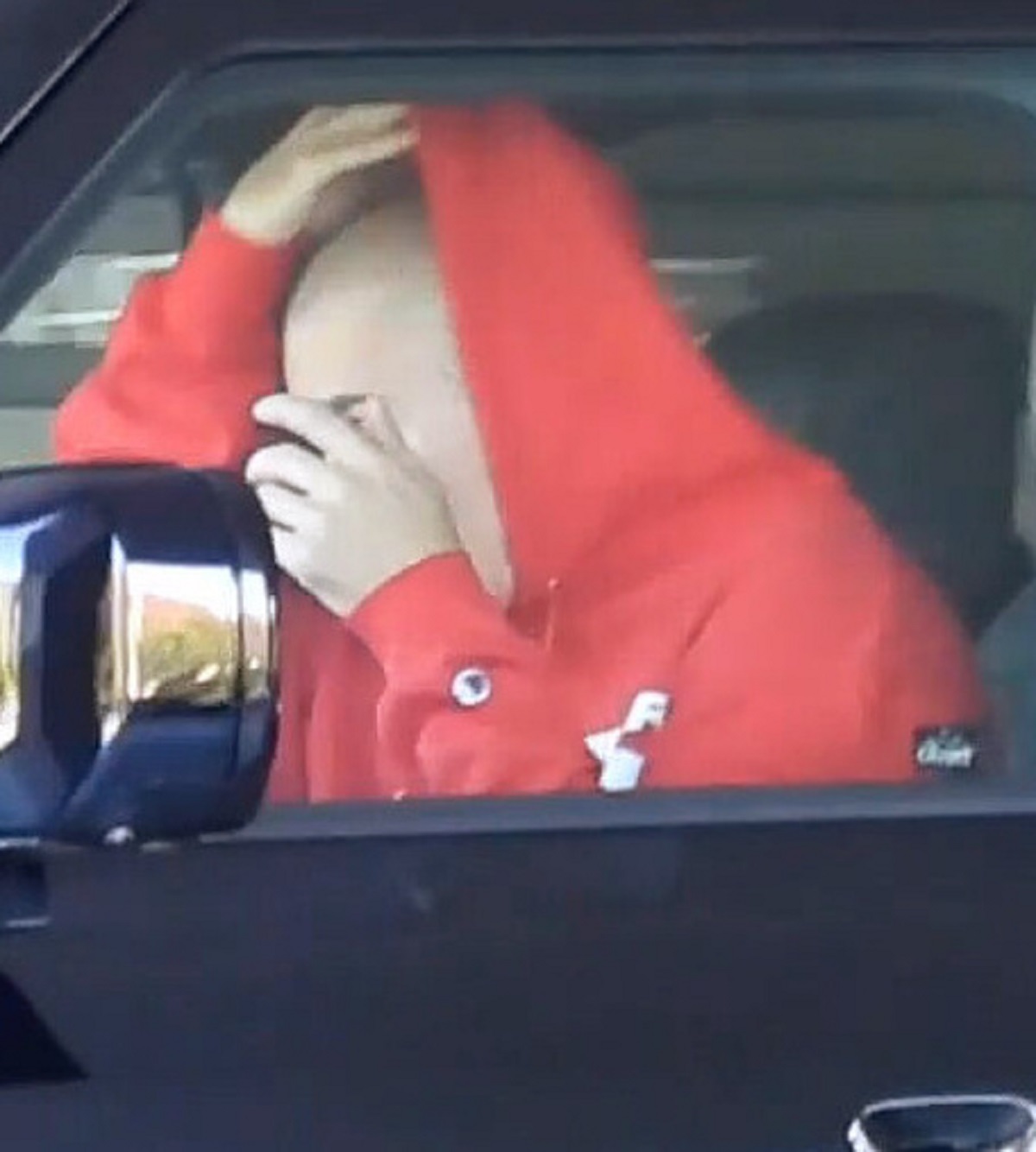 Justin Bieber într-o mașină, purtând un hanorac roșu în timp ce se aranjează în oglindă