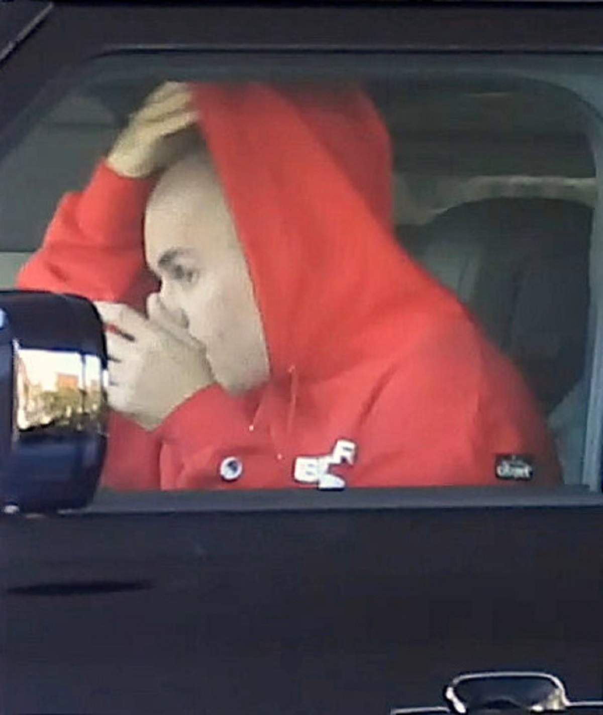 Justin Bieber într-o mașină neagră cu un hanorac roșu în timp ce își aranjează gluga pe capul ras