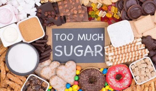 O masă plină cu dulciuri, ciocolată, gogoși, bomboane și alte alimente dulci în mijlocul cărora se află o tăbliță pe care scrie prea mult zahăr