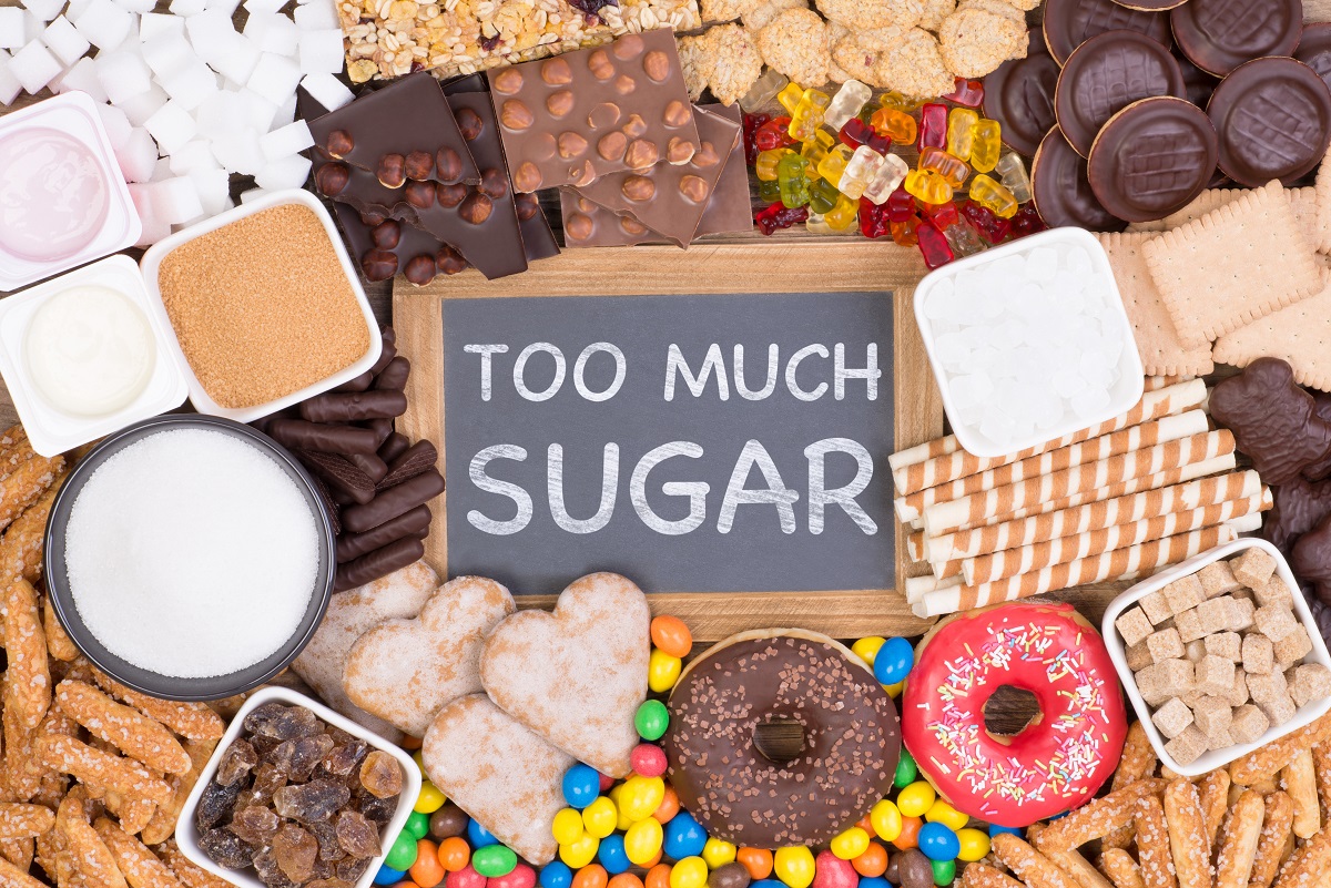 O masă plină cu dulciuri, ciocolată, gogoși, bomboane și alte alimente dulci în mijlocul cărora se află o tăbliță pe care scrie prea mult zahăr