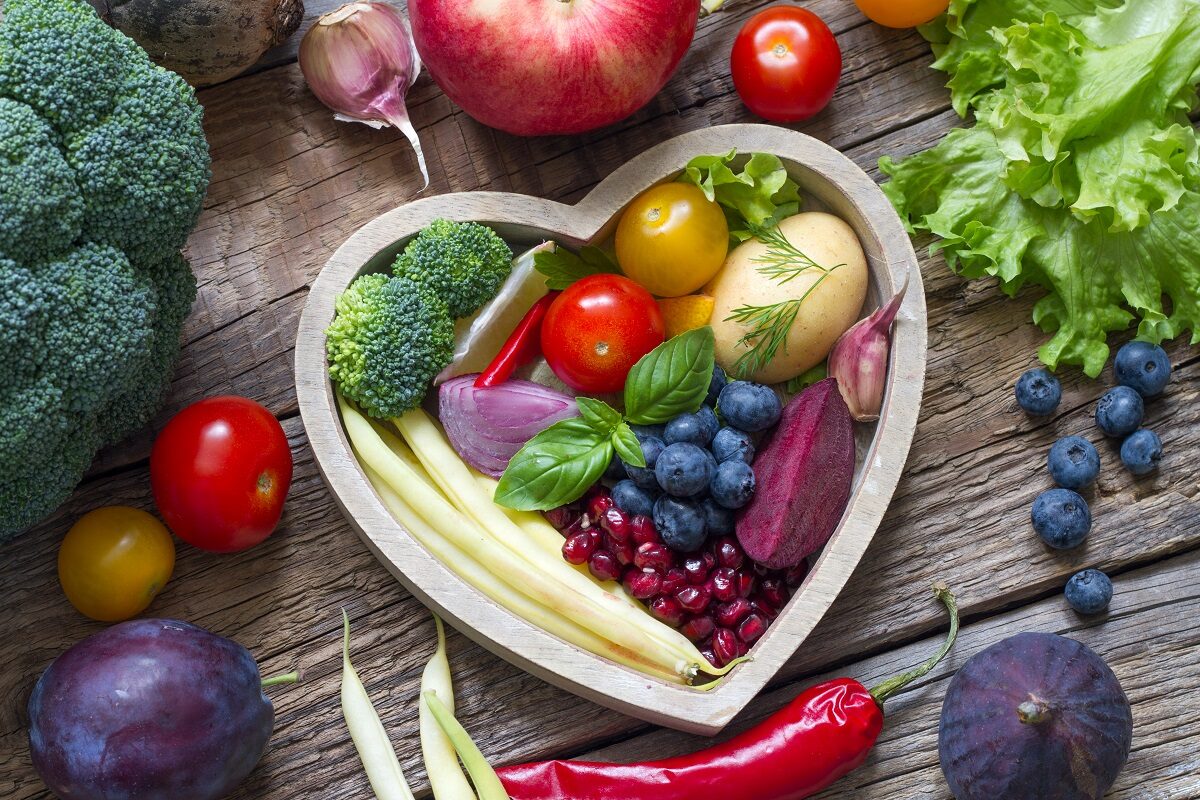 O masă de lemn pe care se află fructe și legume puse într-un bol în formă de inimă pentru a reprezenta una din cele patru metode sănătoase pentru scăderea colesterolului după vârsta de 50 de ani