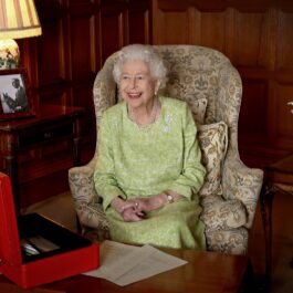 Regina Elisabeta într-o rochie verde în timp ce zâmbește la camerele de fotografiat și stă într-un scaun la Palatul Buckingham