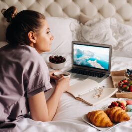 O femeie frumoasă care stă într-un pat în fața unui laptop în timp ce are alături de ea un bol cu fructe de pădure și două cornuri
