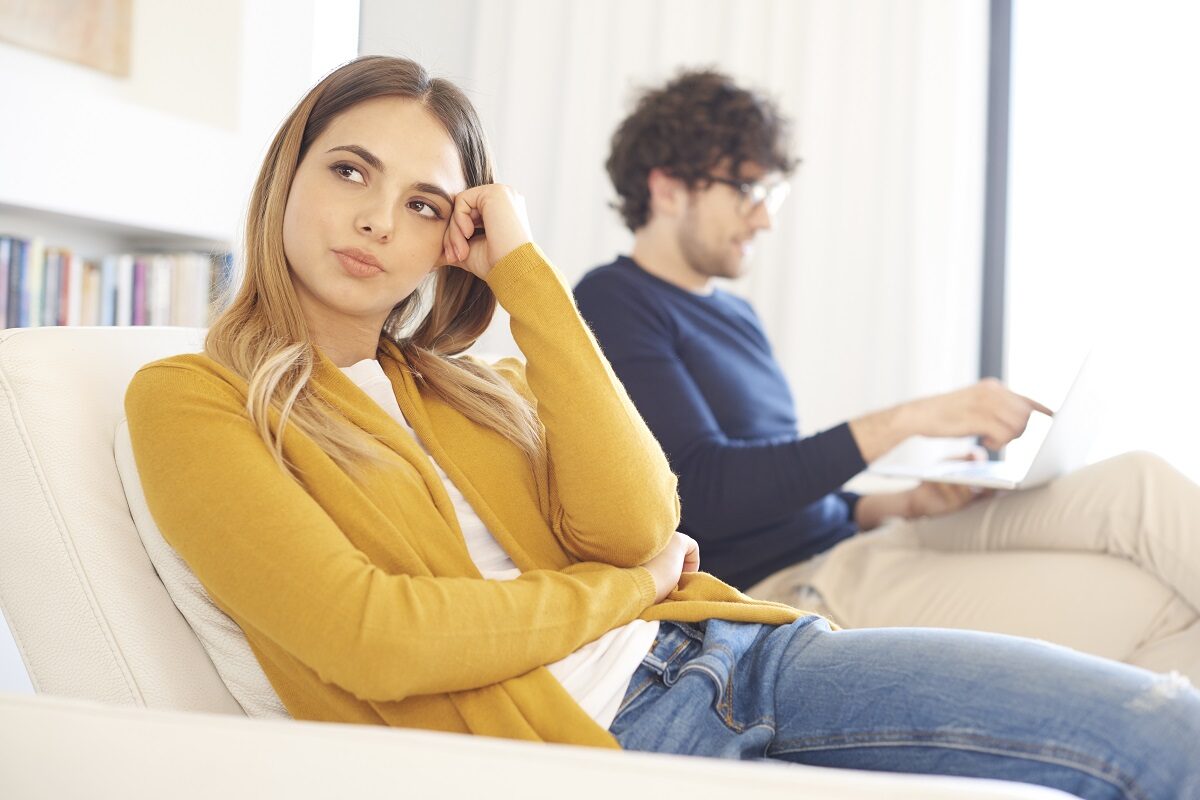O femeie care poată un pulover galben în timp ce stă pe canapea alături de iubitul său și se întreabă dacă ar trebui să renunțe la relație