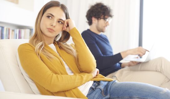 O femeie care poată un pulover galben în timp ce stă pe canapea alături de iubitul său și se întreabă dacă ar trebui să renunțe la relație