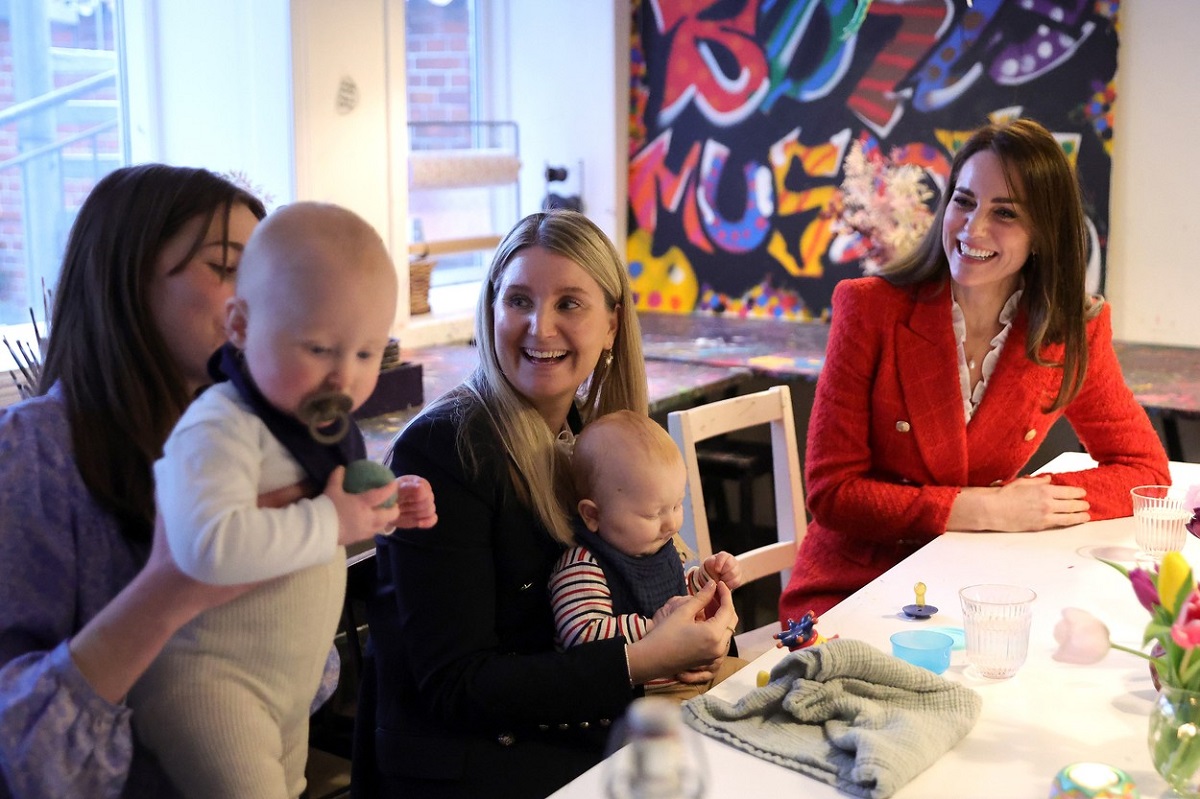 Kate Middleton într-un sacou roșu în timp ce stă de vorbă cu două mămici care își țin bebelușii în brațe în cadrul turului oficial din Danemarca