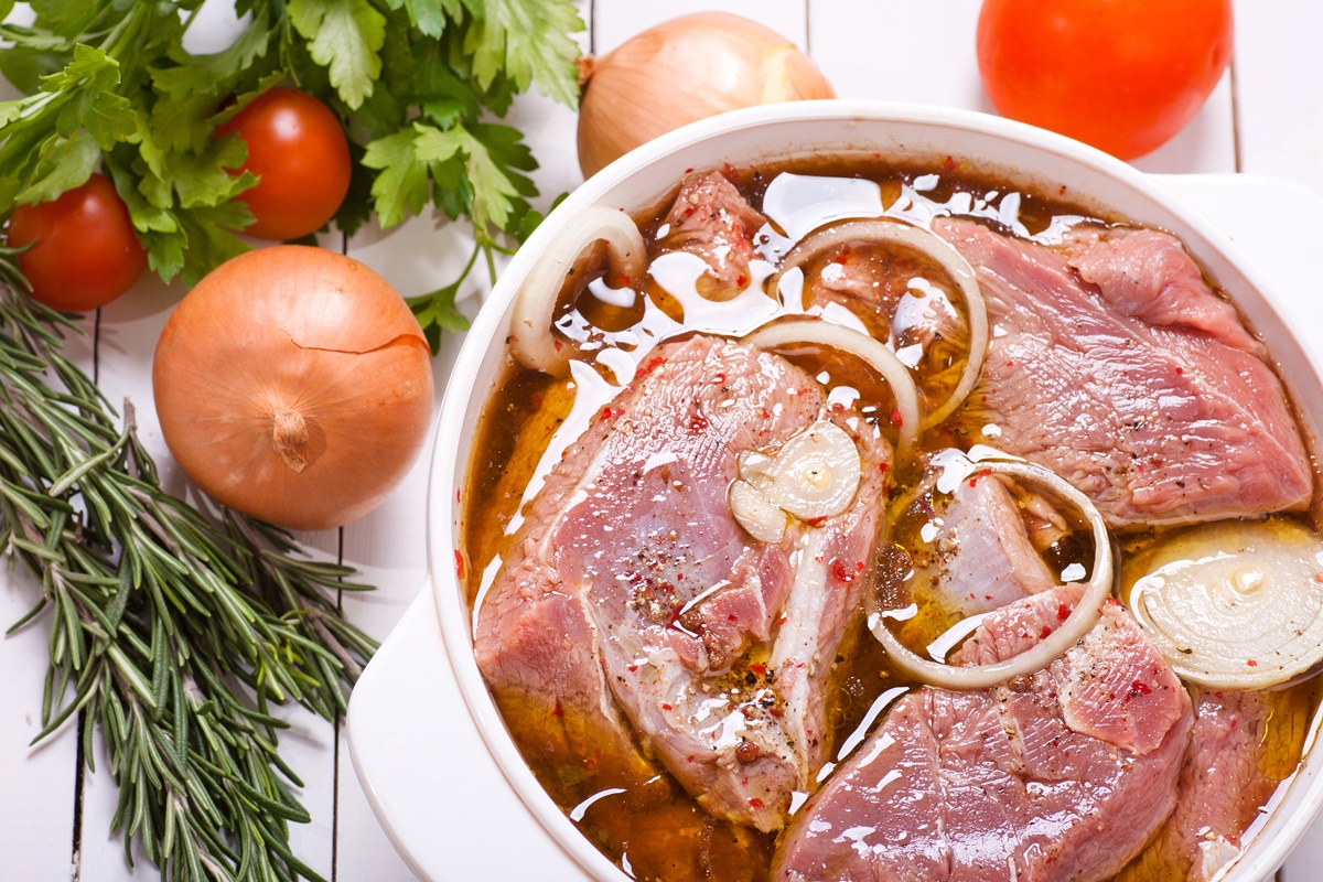 Carne de porc în marinadă într-un bol alb, alături de ceapă, roșii și verdeață, un exemplu despre cum marinezi carnea de porc pentru o friptura cu gust irezistibil