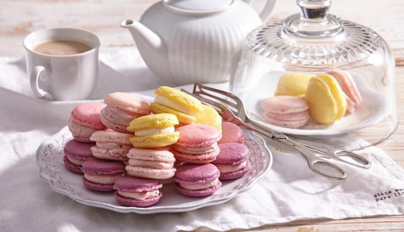 Macarons cu aromă de fructe pe o farfurie și în borcan, alături de o ceașcă cu cafea și cafetieră