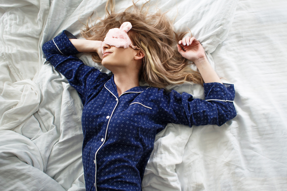 O femeie care doarme cu masca pe față, în pat cu lenjerie deschisă la culoare, în pijamale din mătase