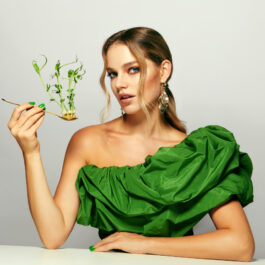 O femeie blondă, în rochie verde, ține în mâna dreptă o lingură, în care se află niște plante verzi.