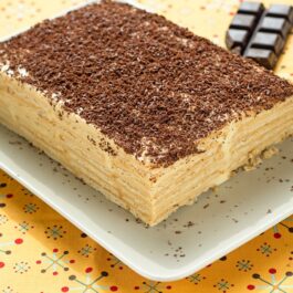 Prăjitură fără coacere cu budincă de vanilie și biscuiți, decorată cu ciocolată răzuită, pe un platou alb