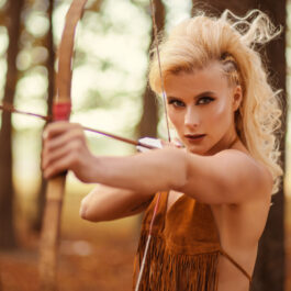 O fată în pădure ține un arc cu săgeți în mână, îndreptat spre o țintă precisă.