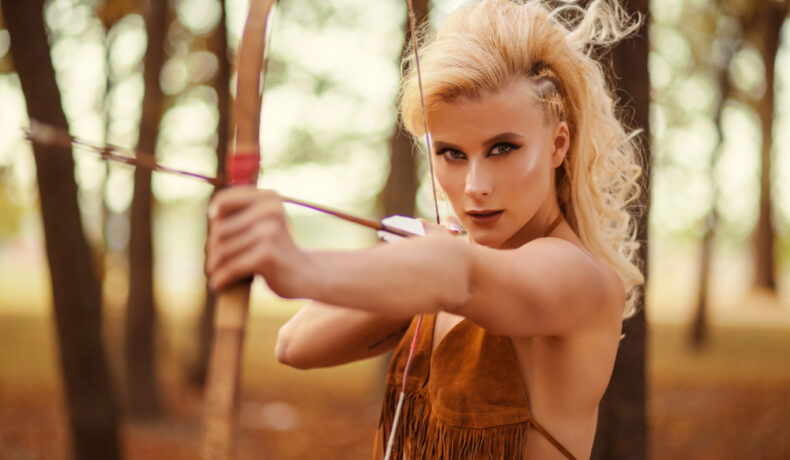 O fată în pădure ține un arc cu săgeți în mână, îndreptat spre o țintă precisă.