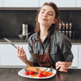 O femeie frumoasă care stă în fața unui blat de bucătărie din lem în timp ce mănâncă dintr-o farfurie pe care se află somon și roșii