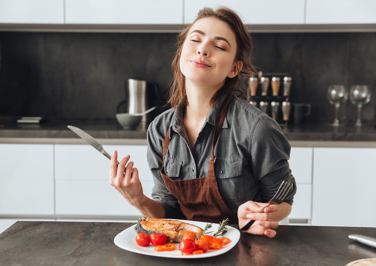 O femeie frumoasă care stă în fața unui blat de bucătărie din lem în timp ce mănâncă dintr-o farfurie pe care se află somon și roșii