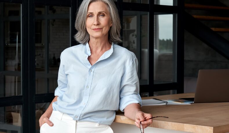 O femeie frumoasă cu părul grizonat care poartă o cămașă albastră și o pereche de pantaloni albi în timp ce se sprijină de un birou pentru a reprezenta una din femeile în vârstă care au adoptat obiceiuri sănătoase pentru viața lor după 60 de ani