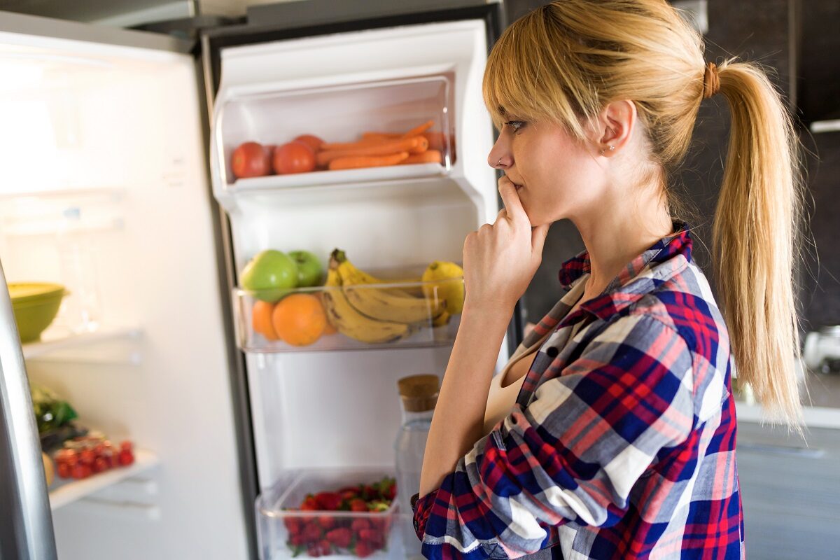 O femeie blondă care poartă o cămașă în carouri și stă în fața unui frigider încercând să aleagă un aliment sănătos