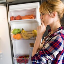 O femeie blondă care poartă o cămașă în carouri și stă în fața unui frigider încercând să aleagă un aliment sănătos