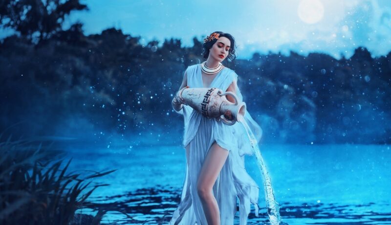O femeie frumoasă îmbrăcată într-o rochie alba care stă pe o stâncă și toarnă apă dintr-un ulcior pentru a reprezenta zodia Vărsător