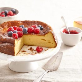 Basque burnt cheesecake secționat pe un platou alb cu picior, alături de un bol cu fructe și un bol cu dulceață de fructe