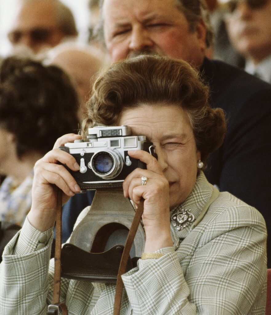Regina Elisabeta cu un aparat de fotografiat în mână în timp ce își lasă la vedere inelul de logodnă primit de la Prințul Philip